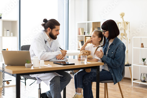 Caucasian male doctor examines Caucasian young girl coughing in clinic, writes medical prescription while Caucasian mother comforts girl.