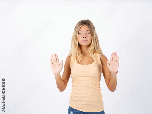 Serious Beautiful teen girl wearing sleeveless t-shirt pulls palms towards camera, makes stop gesture, asks to control your emotions and not be nervous photo