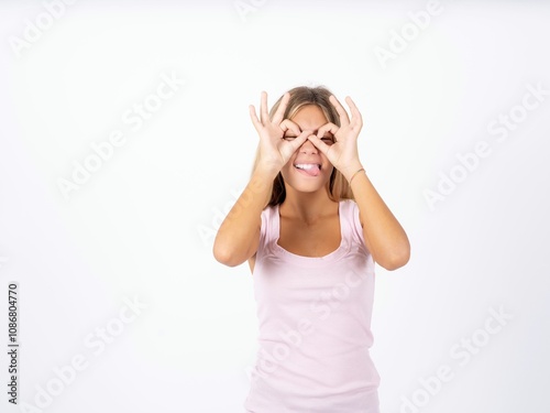 Beautiful teen girl wearing sleeveless t-shirt doing ok gesture like binoculars sticking tongue out, eyes looking through fingers. Crazy expression.