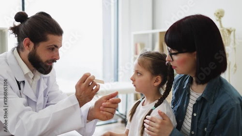 Caucasian male doctor examines young Caucasian girl's throat using a wooden tongue depressor. Girl sits calmly in medical office setting, showing a trustful patient-doctor interaction.