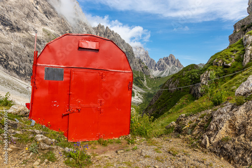 Alpine bivaco mountain hut shelter located in rugged alpine terrain. Red bivacco shelter in Dolomites. Compact structure against stunning landscapes, surrounded by peaks, valleys, and nature. photo