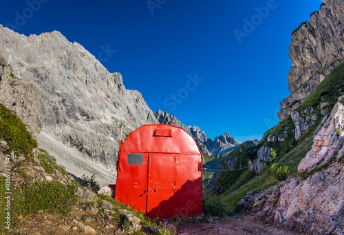 Alpine bivaco mountain hut shelter located in rugged alpine terrain. Red bivacco shelter in Dolomites. Compact structure against stunning landscapes, surrounded by peaks, valleys, and nature. photo