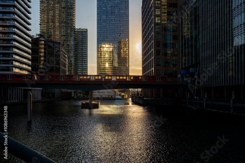 In a stunning contrast of light and shadow, a vibrant red train crosses a bridge over a tranquil waterway, backlit by a radiant golden sunset in the city in London UK photo