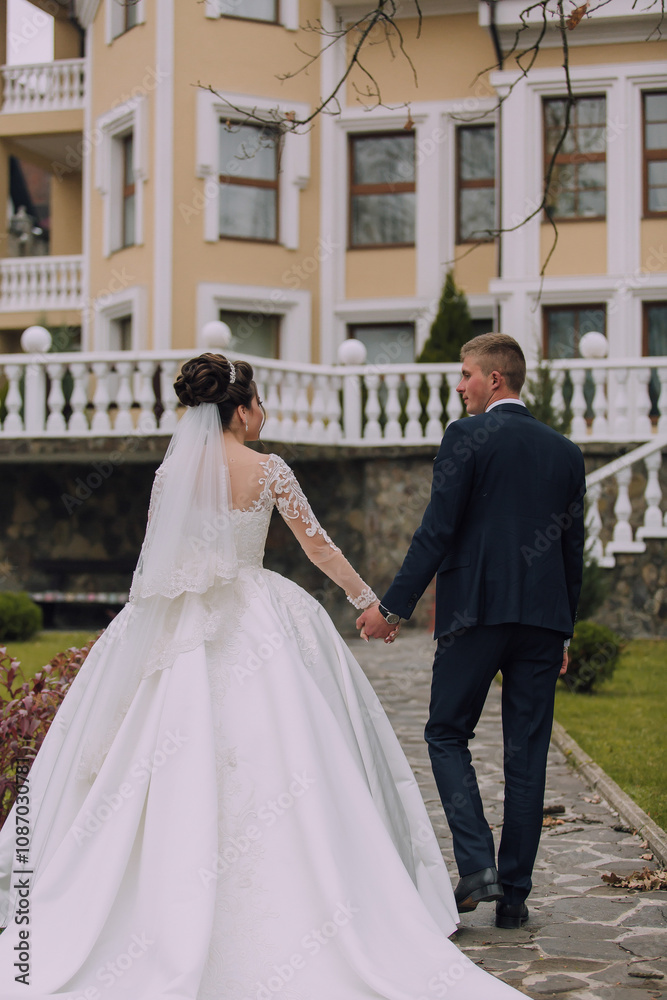 A bride and groom are walking down a path in front of a yellow building. The bride is wearing a white dress and a veil, while the groom is wearing a suit. They are holding hands and appear to be happy A bride and groom are walking down a path in front of a yellow building. The bride is wearing a white dress and a veil, while the groom is wearing a suit. They are holding hands and appear to be happy
