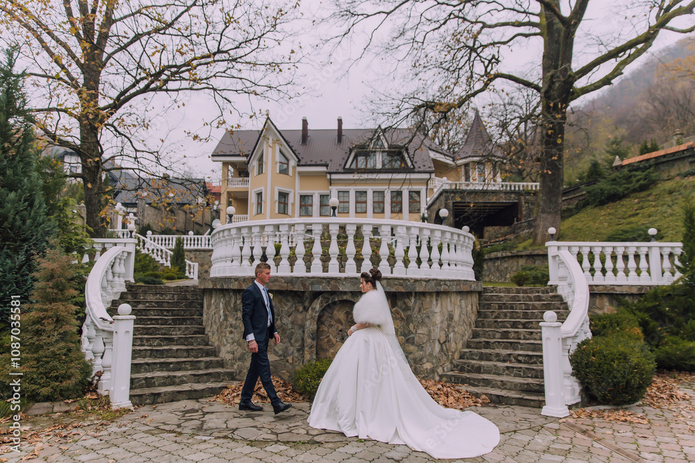 Obraz premium A bride and groom are walking down a set of stairs in front of a yellow house. The bride is wearing a white gown and the groom is wearing a suit. The scene is romantic and intimate