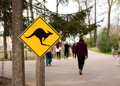 A warning road sign shows the shape of a kangaroo in a park, with people passing by in a blur. photo