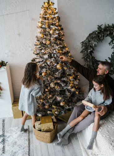 A heartwarming Christmas scene: A father helps his two daughters decorate a beautifully lit Christmas tree. The family s joy and festive spirit are contagious photo