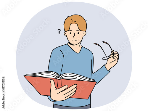 Man with book takes off glasses, trying to make out text in textbook and getting upset because of deterioration of vision. Guy is experiencing vision problems associated with excessive eye strain.