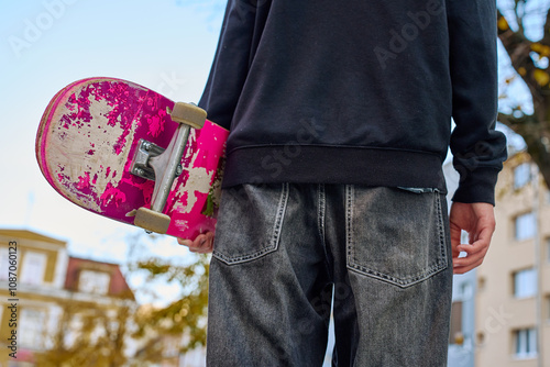 Skateboarder in baggy jeans holding scratched skateboard walking at city street. Teenager is skateboarding. Concept of youth subculture