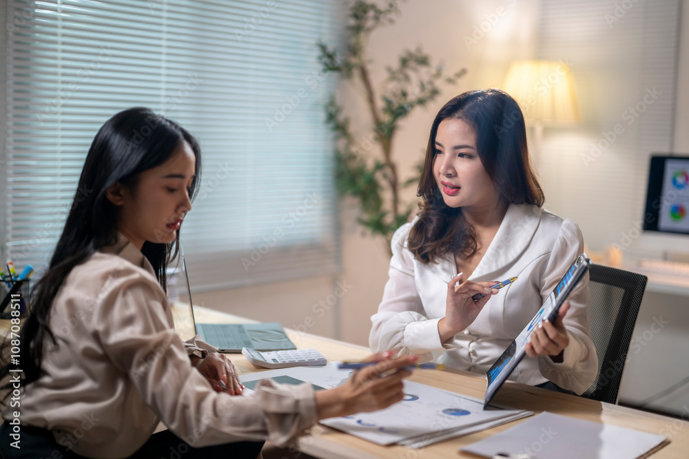 Two women are sitting at a desk, one of them holding a tablet Two women are sitting at a desk, one of them holding a tablet