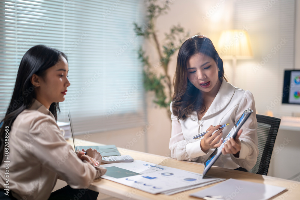 Two women are sitting at a desk, one of them is pointing at a tablet Two women are sitting at a desk, one of them is pointing at a tablet