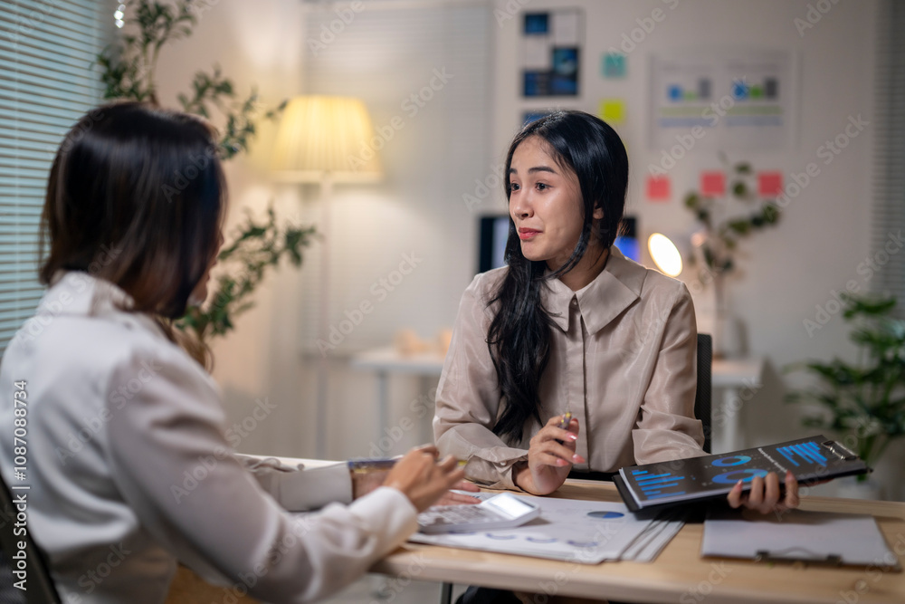 Two women are sitting at a desk, talking to each other Two women are sitting at a desk, talking to each other
