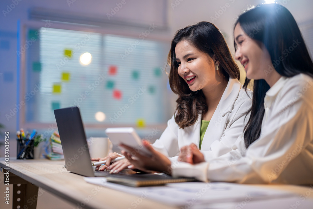 Two women are sitting at a desk with a laptop and a cell phone Two women are sitting at a desk with a laptop and a cell phone