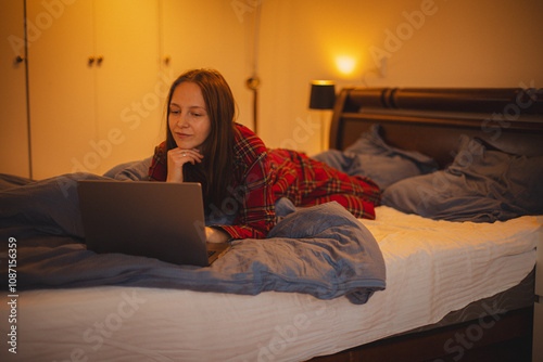 young woman in festive plaid pajamas lies in bed in a cozy bedroom, peacefully browsing her laptop photo