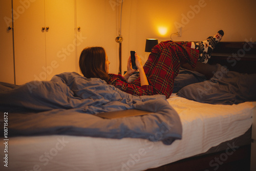 young woman in plaid pajamas lies in bed in a cozy bedroom, peacefully browsing her phone. Perfect for concepts of mindfulness, Christmas mornings, and holiday relaxation.  photo