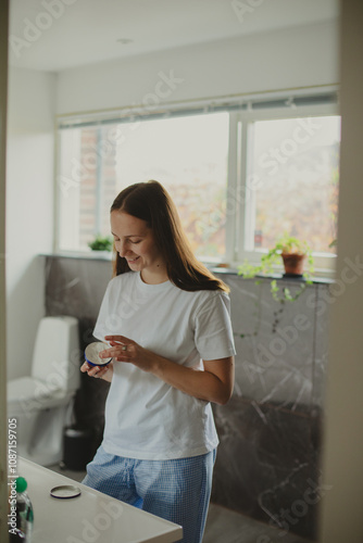 Happy Woman Applying Face Cream Moisturizing And Caring For Skin Smiling To Her Reflection In Mirror Standing In Modern Bathroom At Home. Facial Skincare And Pampering. Selective Focus photo