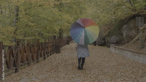 Woman with rainbow colored umbrella walking among colorful leaves in autumn