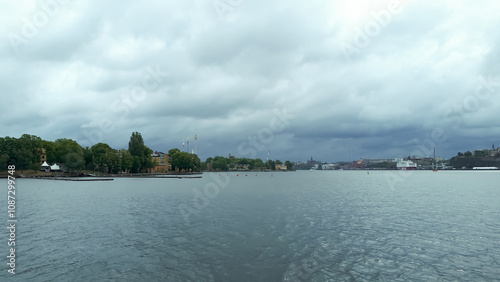 Overcast sky over a serene waterfront with lush greenery and distant cityscape.