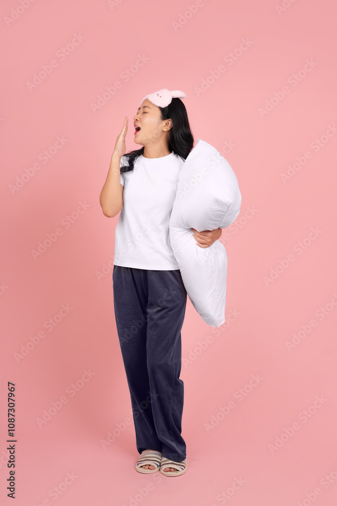 Full body shot of tired asian woman in white nightgown and eye mask hugging pillow and yawning covering mouth with hand, standing on pink background Full body shot of tired asian woman in white nightgown and eye mask hugging pillow and yawning covering mouth with hand, standing on pink background