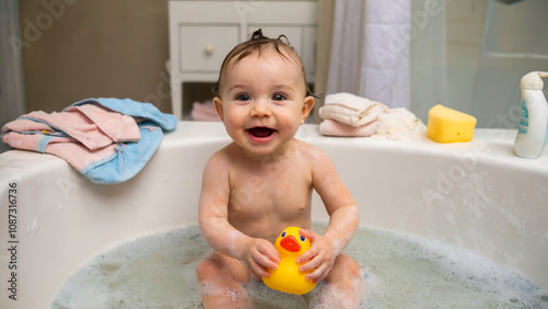 A happy baby in a bubble-filled bathtub, enjoying playtime with a yellow rubber duck, surrounded by towels and baby care items. photo