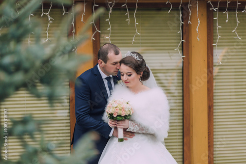 A bride and groom are hugging and kissing each other. The bride is wearing a white fur stole and a bouquet of flowers photo