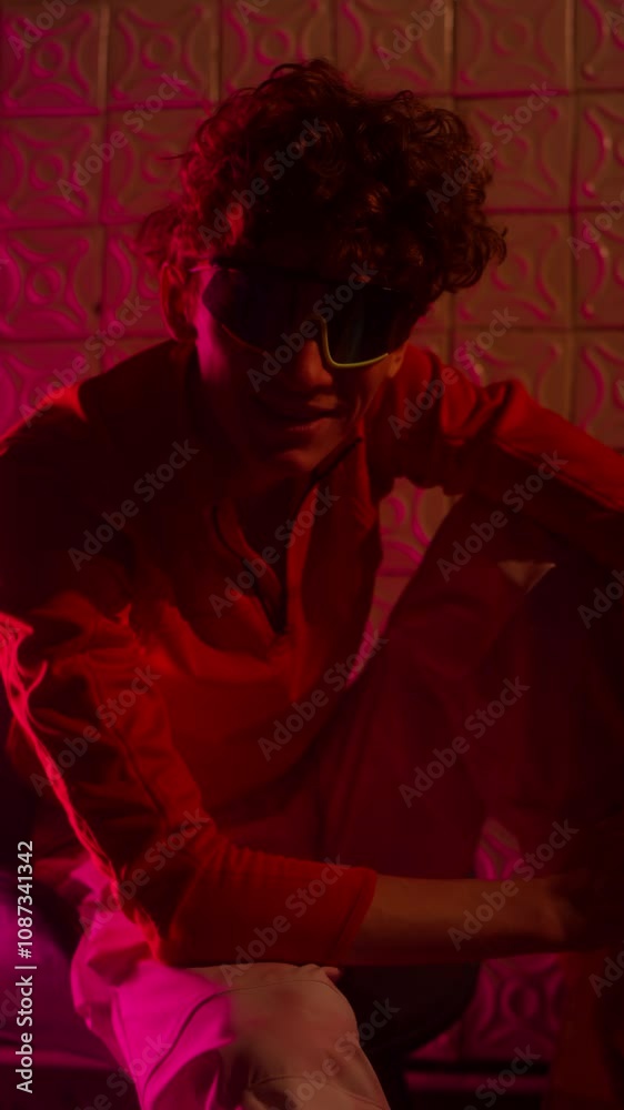 Vertical portrait of curly-haired gen Z guy with glasses leaning towards camera and smiling while sitting on chair in club with neon light Vertical portrait of curly-haired gen Z guy with glasses leaning towards camera and smiling while sitting on chair in club with neon light