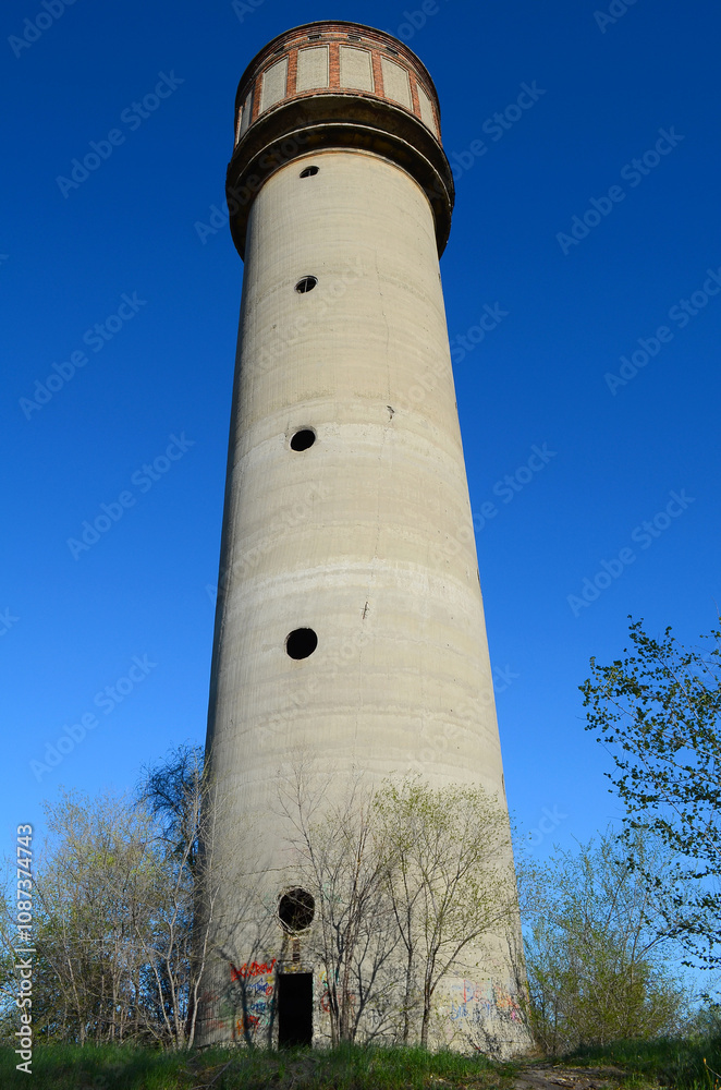 An old, abandoned concrete water tower with graffiti at its base stands tall against a clear blue sky, surrounded by trees. An old, abandoned concrete water tower with graffiti at its base stands tall against a clear blue sky, surrounded by trees.