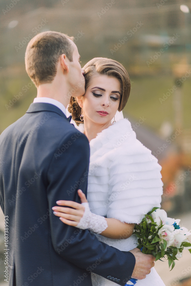 A bride and groom are embracing each other, with the bride wearing a white fur stole. The bride is holding a bouquet of flowers in her right hand. The scene is intimate and romantic A bride and groom are embracing each other, with the bride wearing a white fur stole. The bride is holding a bouquet of flowers in her right hand. The scene is intimate and romantic