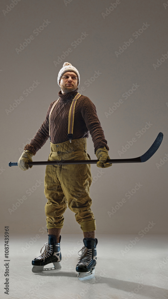 Full-length portrait of man, hockey player in sweater, pants with suspenders and knitted hat, standing with stick in grey studio background. Concept of sport, game, tournament, action, competition Full-length portrait of man, hockey player in sweater, pants with suspenders and knitted hat, standing with stick in grey studio background. Concept of sport, game, tournament, action, competition