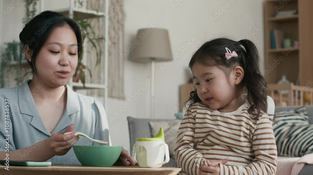 Young Asian woman offering her cute little daughter porridge for breakfast and laughing at her when naughty girl turning her head away and refusing to eat homemade meal Young Asian woman offering her cute little daughter porridge for breakfast and laughing at her when naughty girl turning her head away and refusing to eat homemade meal