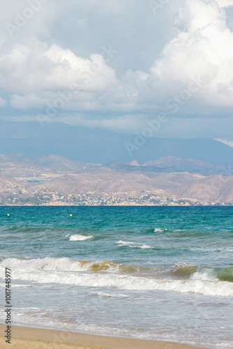 Gentle waves lapping against the sandy beach, framed by a distant mountainous coastline and a cloudy sky. Alicante, Spain. 