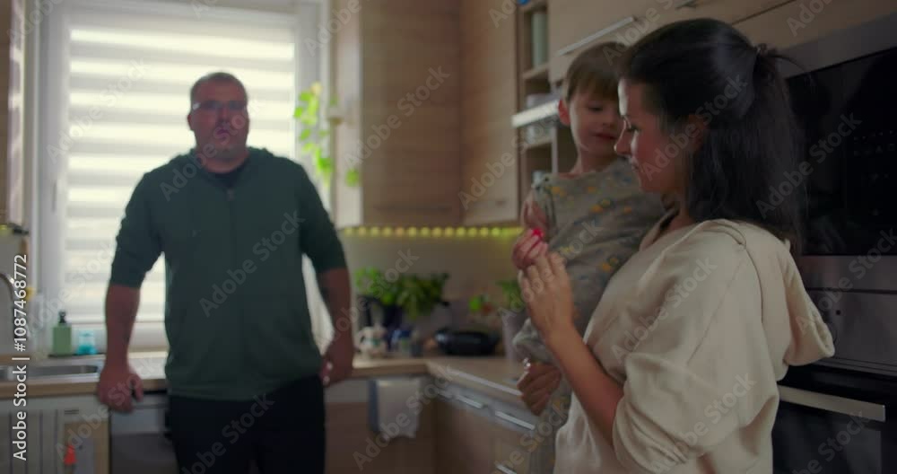 Mother holding her child and playfully interacting, while a man stands in the background in a bright, modern kitchen with wooden interior elements