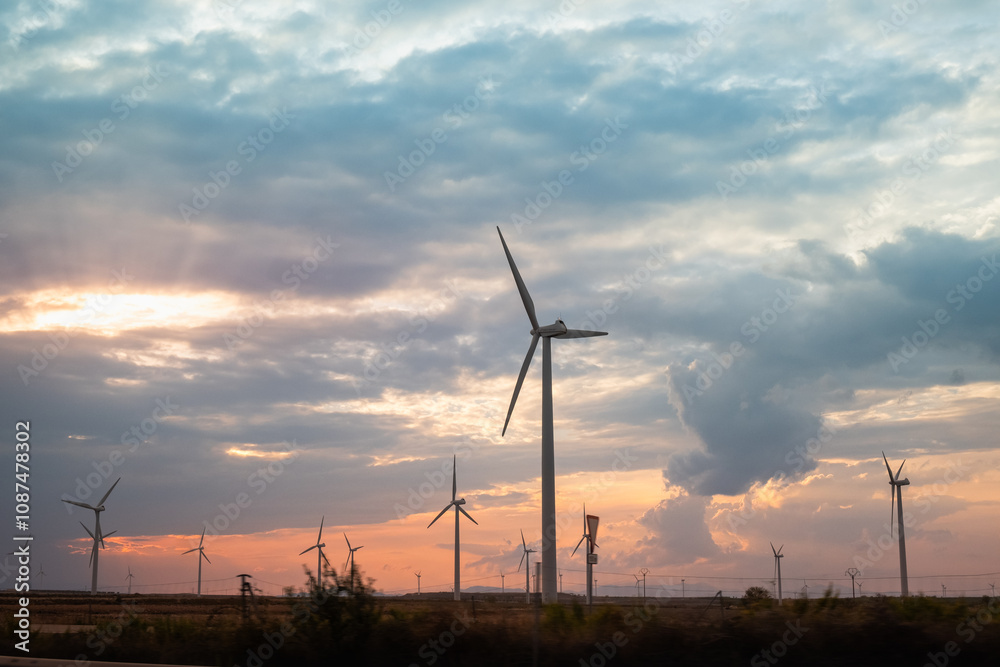 Obraz premium side view of a large wind farm with a nice stormy sky at sunset. The windmills or generators face the sun and the sky is full of clouds