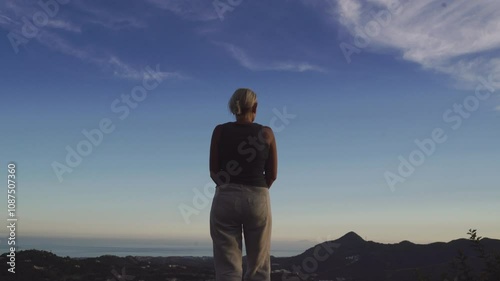 A woman stands on a mountain overlook, gazing at the expansive landscape under a clear blue sky. Captures peaceful solitude and connection with nature