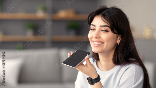 Digital Speaker Application. Portrait of young smiling woman sitting at table, talking on mobile phone, using laptop and virtual voice assistant for online search, to record reminder, dictate message