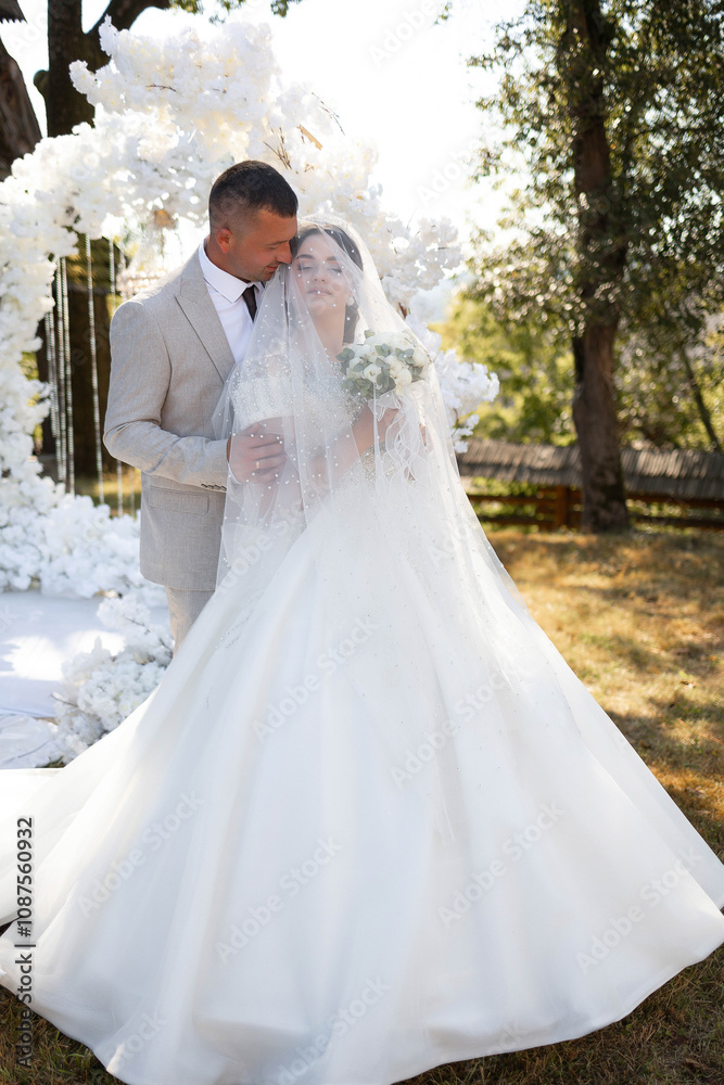 A bride and groom are standing in front of a white archway. The bride is wearing a white dress and the groom is wearing a suit. They are both smiling and hugging each other A bride and groom are standing in front of a white archway. The bride is wearing a white dress and the groom is wearing a suit. They are both smiling and hugging each other
