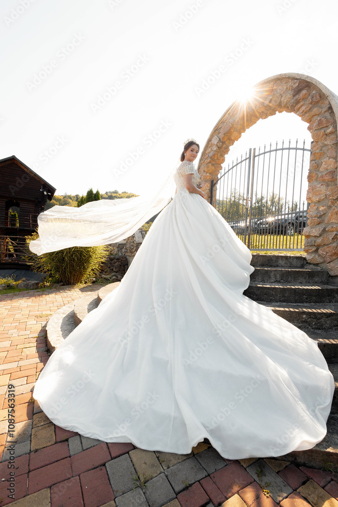 Fototapeta premium A woman in a white dress is posing in front of a stone archway. The dress is long and flowing, and the woman is wearing a tiara. Concept of elegance and sophistication, as the woman's attire
