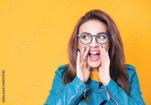 Amused woman yelling, wearing blue leather jacket, isolated on yellow background. Happy girl holding hands near his mouth