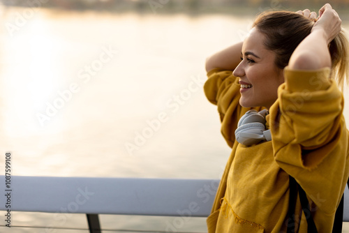 A young woman enjoys a peaceful moment by the waterfront at sunset, basking in the golden light while adjusting her hair with relaxed joy