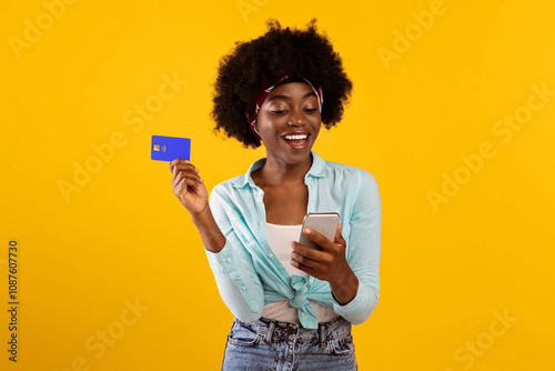 Mobile Shopping Application. Happy African Lady Using Smartphone Holding Credit Card Paying Online Buying Clothes In Shopper App Standing Over Pink Background. Studio Shot