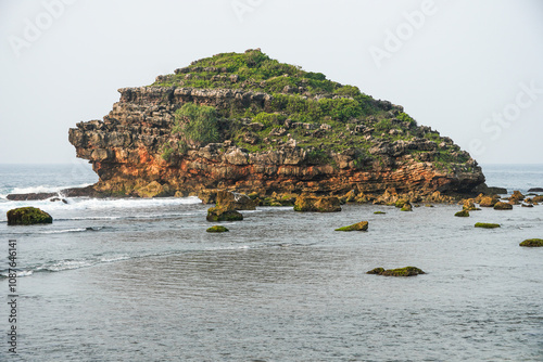 Rocky headland with crashing waves during the day. Cape with cliffs by the sea and green plants on it at tropical island in Watu Karung Beach, Pacitan, East Java, Indonesia photo