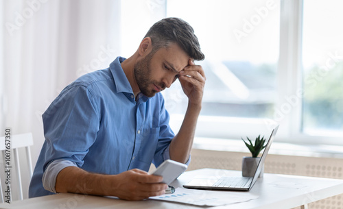Worried male freelancer looking at smartphone screen while working at home office, received bad news, got scam message, thoughtful man touching forehead with concerned face expression, copy space