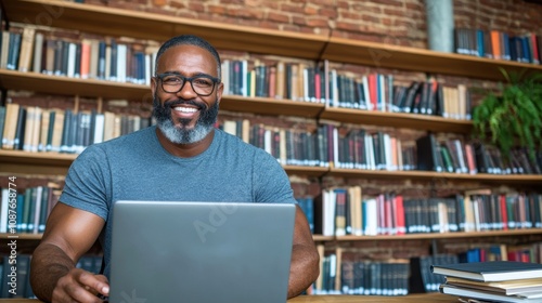 A man with a beard and glasses sits at a table, engaging with his laptop in a library brimming with a vast collection of books. The warm atmosphere encourages productivity