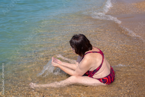 Mature woman in swimsuit, rear view, sits on seashore and splashes in water. Vacation at sea for pensioners