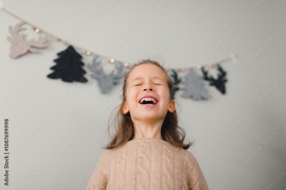 A little girl is laughing in front of a Christmas garland with fir trees and reindeer. Merry Christmas and Happy New Year concept A little girl is laughing in front of a Christmas garland with fir trees and reindeer. Merry Christmas and Happy New Year concept