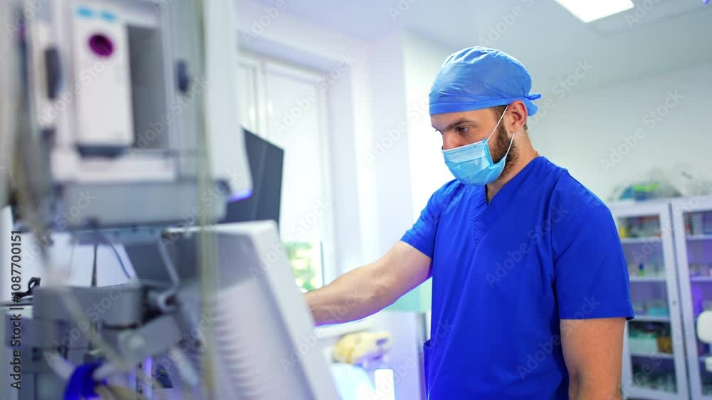 Bearded medic in blue uniform stands at the medical equipment. Doctor pressing the keys on the machine during operation. Bearded medic in blue uniform stands at the medical equipment. Doctor pressing the keys on the machine during operation.