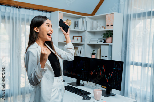 Successful wealthy with extremely smiling in young Asian businesswoman earning high profit raising fist up with standing jump, holding smartphone in exchange stock market at modern office. Stratagem. photo