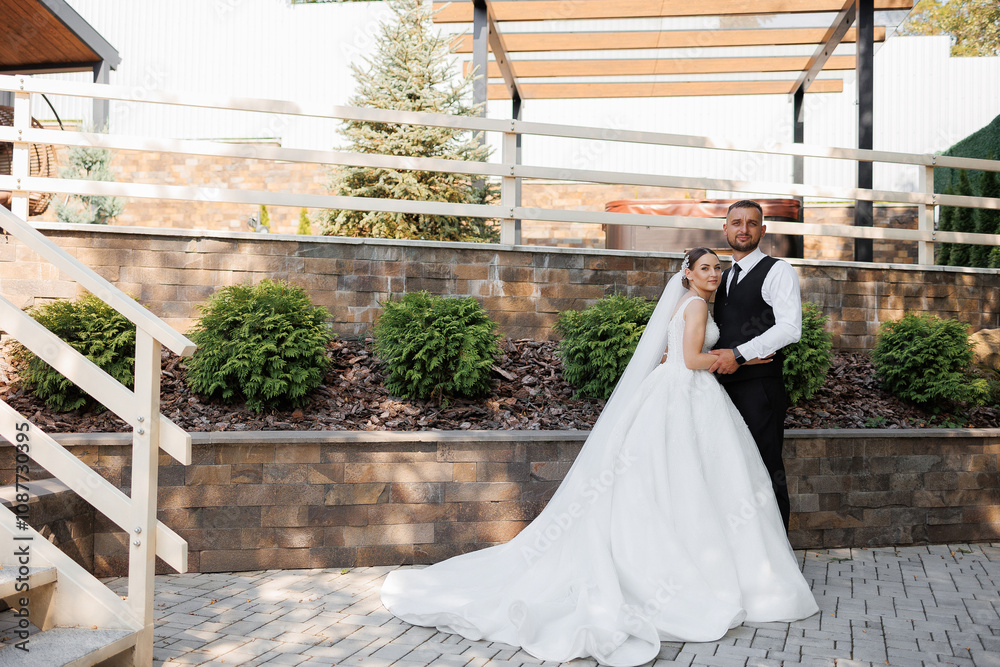 Fototapeta premium A bride and groom are posing for a picture in front of a brick wall. The bride is wearing a white dress and the groom is wearing a black vest. Scene is happy and celebratory