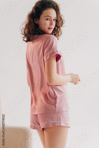Curly girl in pink silk pajama. Beautiful young woman near armchair on white background.