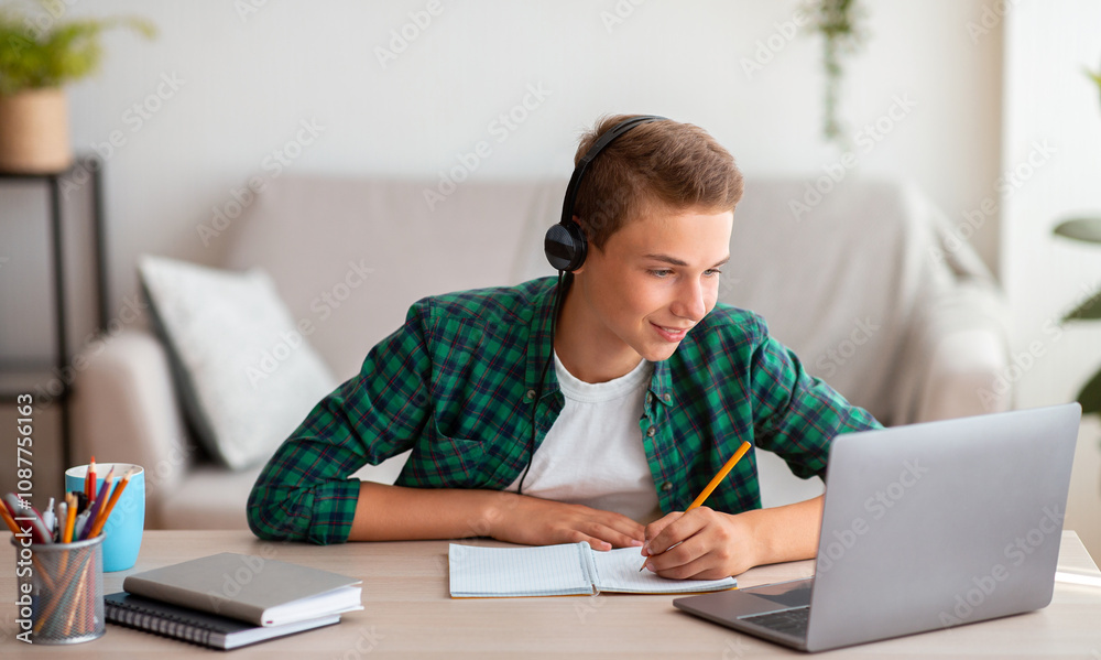 Joyful teen guy writing at notebook and looking at laptop screen, having lesson online, home interior, free space Joyful teen guy writing at notebook and looking at laptop screen, having lesson online, home interior, free space